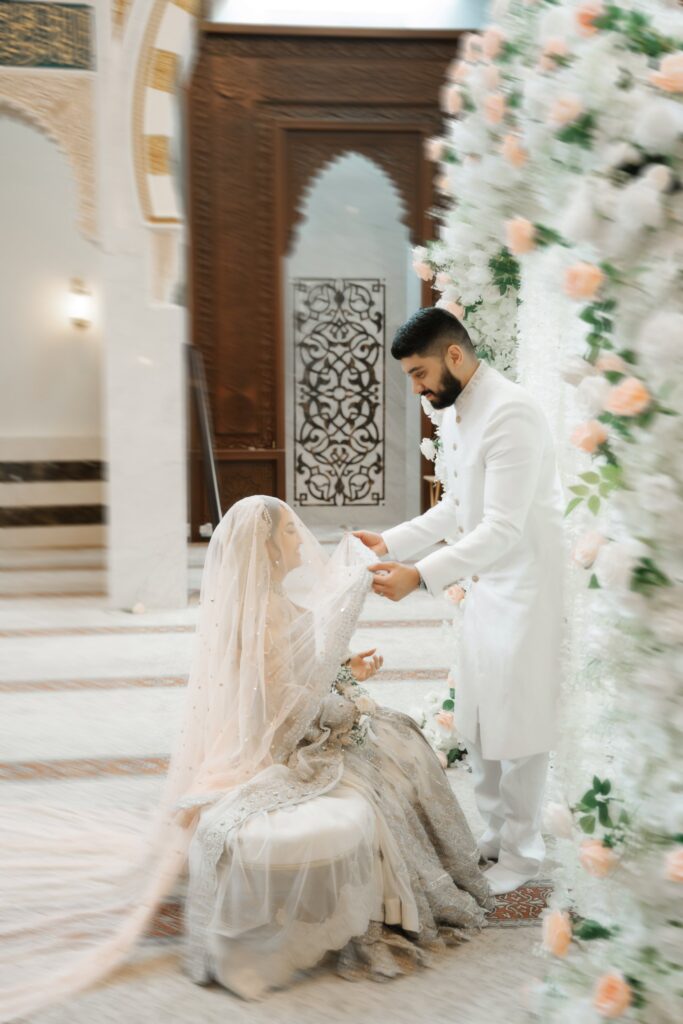 Groom lifting the bride’s veil beside a white and peach floral wall at their nikah in Sanford, Florida