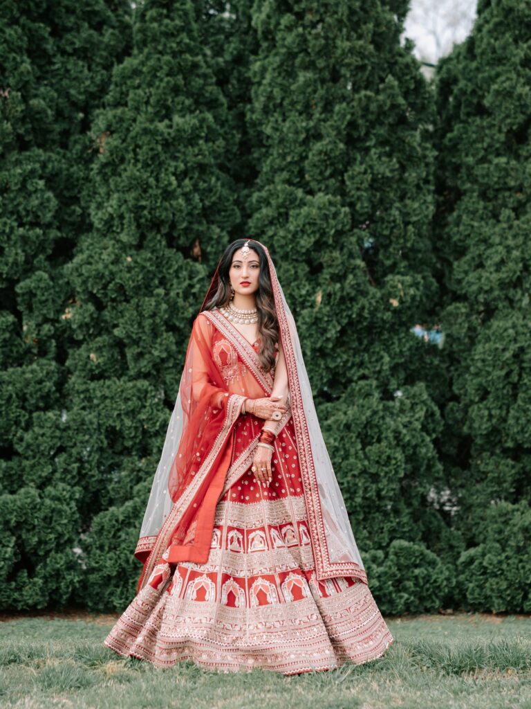 Priya & Dillan’s Gujarati Wedding | Indian Wedding Photographer Atlanta 12 Gujarati bride in a red lehenga with a sheer red dupatta draped over her head in front of evergreen trees