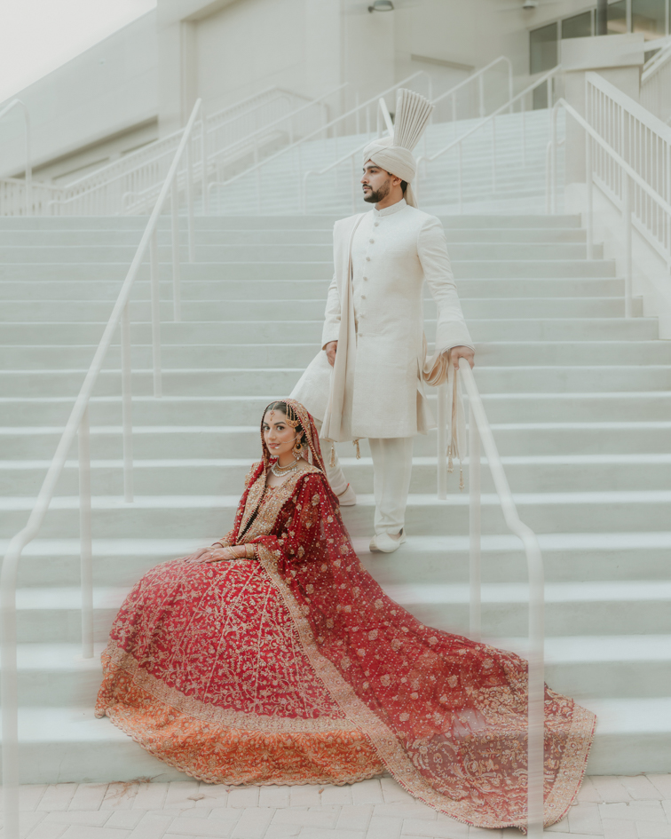 Indian Wedding Venues in Florida 3 South Asian wedding photographer Orlando: bride seated on white stairs with her red lehenga spread out, groom standing behind