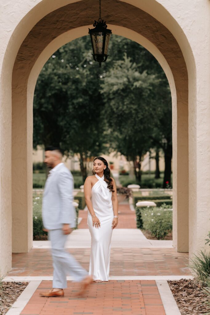 Rahul & Nirali’s Romantic Rollins College Engagement Session 7 Woman in a white satin halter gown under a stucco arch as her fiancé walks past, Winter Park engagement photos