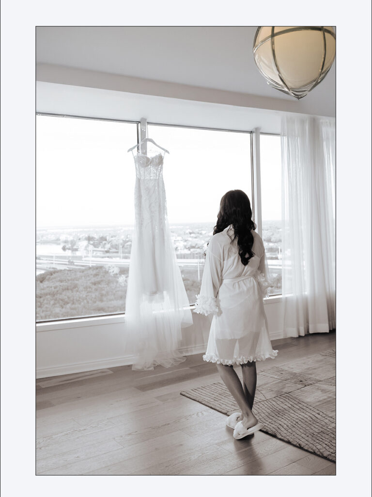 Shobin & Maria’s Malayali Christian Wedding at Grand Hyatt Tampa Bay 1 Malayali bride in a white robe looking out the window at her wedding gown hanging in her suite at Grand Hyatt Tampa Bay