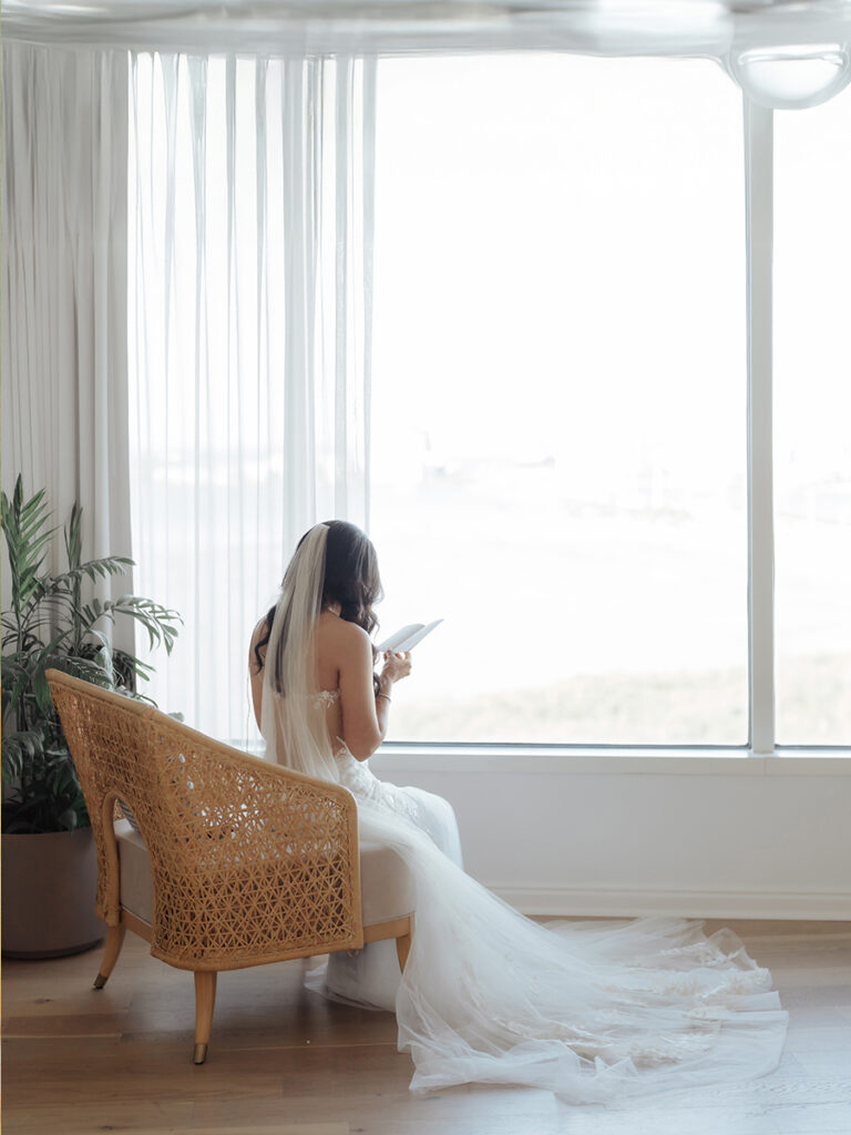 Shobin & Maria’s Malayali Christian Wedding at Grand Hyatt Tampa Bay 3 Bride in a cathedral veil reading a letter in a rattan chair by tall windows overlooking Tampa Bay