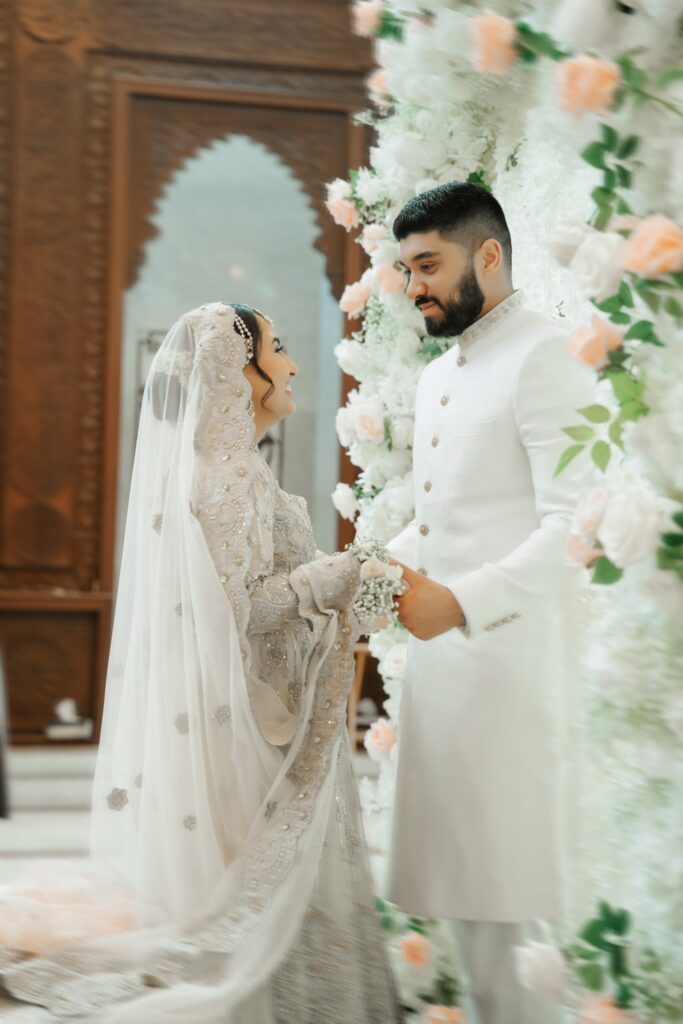 Bride and groom smiling at each other beside a white rose flower wall inside Masjid Al-Hayy